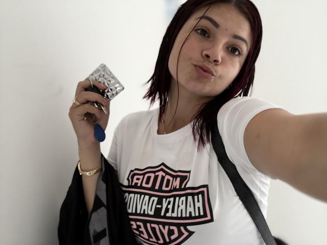 KatherineToro with red hair, white t-shirt, holding condom in right hand, black bag on shoulder, gold bracelet, pouty expression, selfie. Boobs not visible.