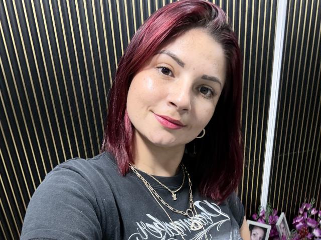 KatherineToro with red hair, wearing a grey shirt and gold necklace, smiling slightly, showing her shiny skin and pink lipstick, with a hoop earring in her left ear.