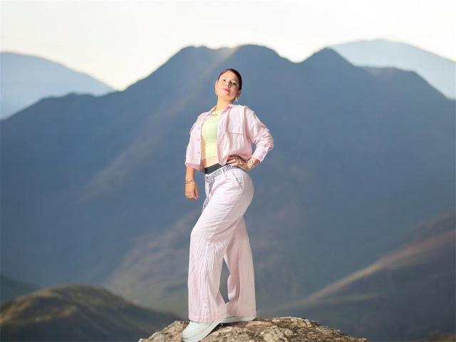 KatherineToro stands on a rock, wearing a pink jacket, white shirt, and pink pants, hands on hips, looking up, with mountains in the background, and a tattoo on her wrist.