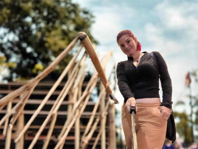 KatherineToro with red hair, black long-sleeve top, tan pants, hands in pockets, holding a golf club, smiling, standing by a wooden bridge, clear blue sky, trees.