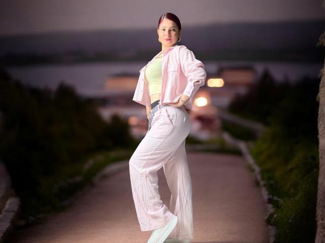 KatherineToro with pale pink jacket, white high-waisted pants, and green top, standing on a path at night, hands in pockets, ass slightly raised, looking confident.