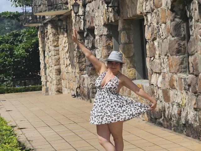 AlessiaRed, in a white floral dress with a deep V-neck, stands on a stone pathway, arms raised, wearing a gray bucket hat, smiling, her ass slightly visible.