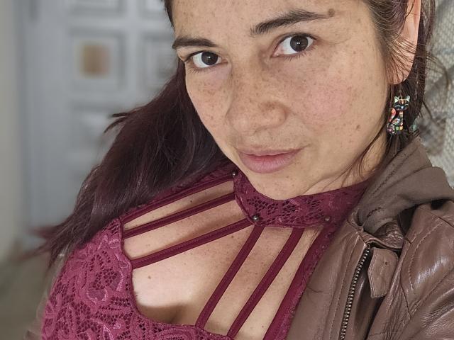 AlessiaRed with freckles, wearing a lace maroon top with strappy design, brown leather jacket, and colorful earrings, looking at the camera with a slight smile.