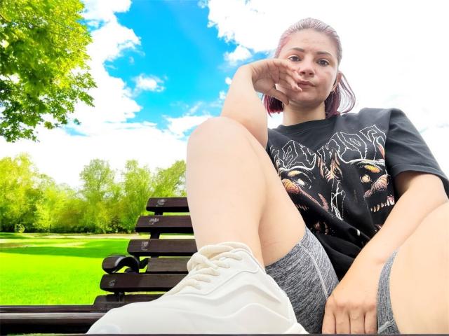 KatherineToro, sitting on a black bench, wearing a black rock t-shirt and gray shorts, with one leg up, pouting, and white sneakers, in a bright, green park.