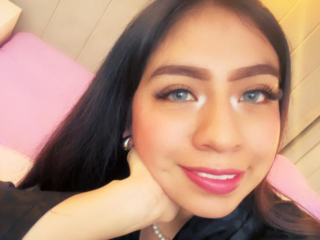ZoeRedds a de longs cheveux noirs, des yeux bleus et du rouge à lèvres rose. Elle sourit, une main sous son menton, mettant en valeur son visage séduisant et ses jolies lèvres roses..