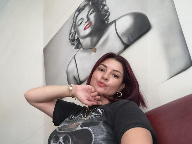 Katherine Toro, con cabello rojo, camiseta negra con estampado de calavera y aros grandes, con la mano cerca de la barbilla, y un cuadro en blanco y negro de Marilyn detrás de ella..