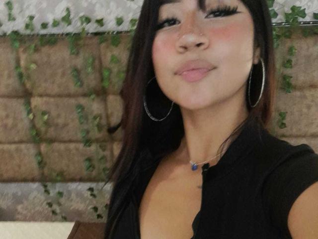 BonnieBond has medium brown skin, black hair, and big hoop earrings. She's wearing a black top with a blue pendant, smiling with pink cheeks and long eyelashes.