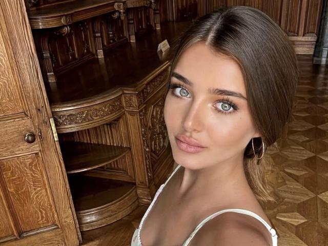 NittaDali hat blaue Augen, braunes Haar im Pferdeschwanz, eine weiße Spitzenbluse und kleine Brüste. Sie trägt goldene Ohrringe im Stecker-Stil und lächelt leicht in die Kamera..