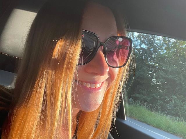 Anna89-hot with long brown hair, wearing black sunglasses, smiling in a car with sunlight on her face, visible reflection in glasses, and a slight tan.