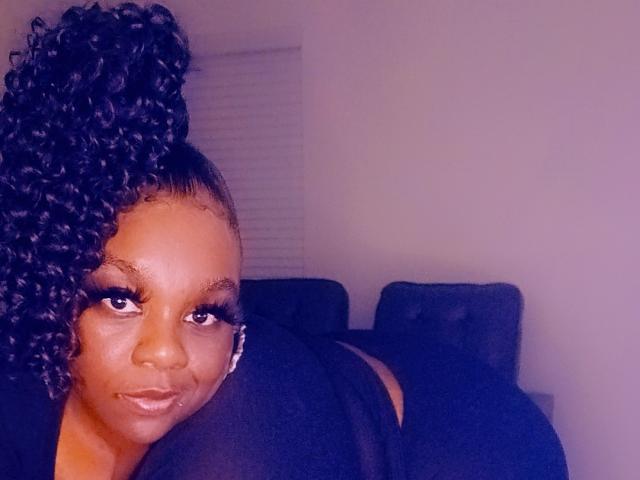 MzThundaBooty with dark curly hair in a high ponytail, looking at the camera with a slight smile, wearing black clothing, showing her ass in the background.