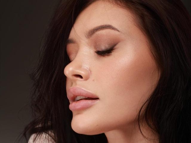 El rostro de MilanaMusee, con los ojos cerrados y los labios rosados ligeramente entreabiertos, muestra una piel suave y clara, y cabello ondulado de color marrón oscuro. Tiene una pequeña marca de nacimiento en su mejilla izquierda..