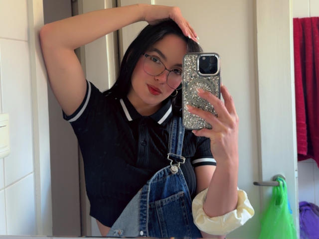 MaiaPark with glasses, black shirt, denim overalls, hand in hair, holding glitter phone, red lipstick, white scrunchie, mirror selfie, smiling, black hair, cute.