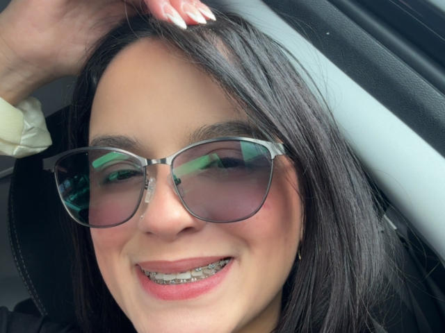 MaiaPark with black hair, wearing reflective sunglasses, smiling with metal braces, white nails, hand in hair, sitting in a car.