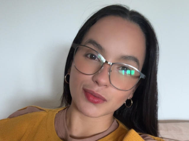 MaiaPark has dark, straight hair, wears glasses with a green reflection, red lipstick, hoop earrings, and a yellow-brown top. Her ass is not visible.
