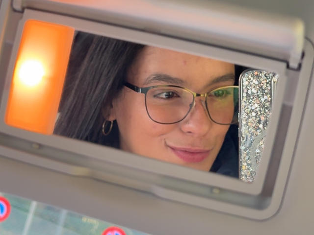 MaiaPark's face in a car mirror, wearing glasses and a gold hoop earring, with a slight smile and a small bruise on her left cheek.
