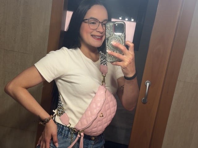 MaiaPark with glasses, smiling, holding phone, pink quilted purse over tits, white shirt, blue jeans, wooden door, mirror selfie, wearing bracelet, dark hair, red lipstick.