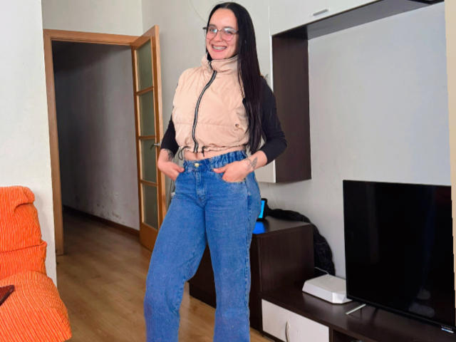 MaiaPark with long black hair, glasses, smiling, wearing a beige zip-up vest, black long-sleeve shirt, and blue high-waisted jeans, hands in pockets, standing in a room.