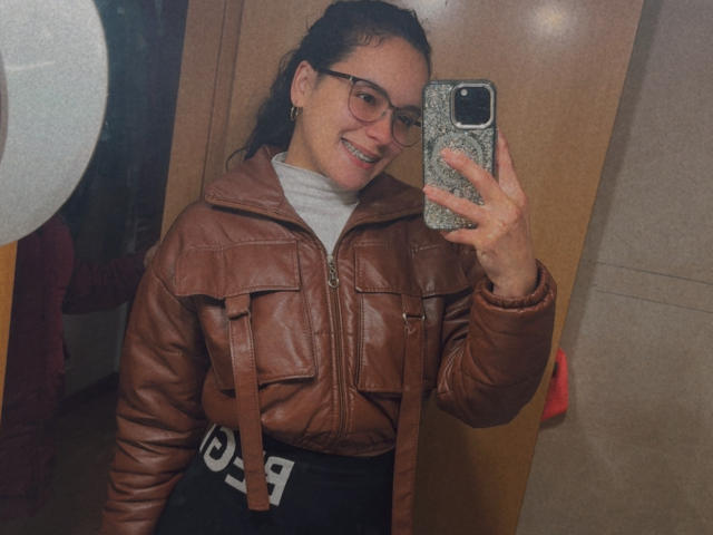 MaiaPark with glasses, brown leather jacket, gray turtleneck, black pants, smiling, holding phone with gray pattern, dark hair in ponytail, mirror selfie.