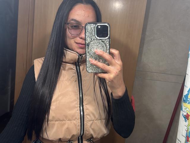 MaiaPark with long black hair, glasses, beige puffy vest, black long-sleeve, holding glitter phone, taking mirror selfie, slight smile, wooden background, visible red scratch on face.