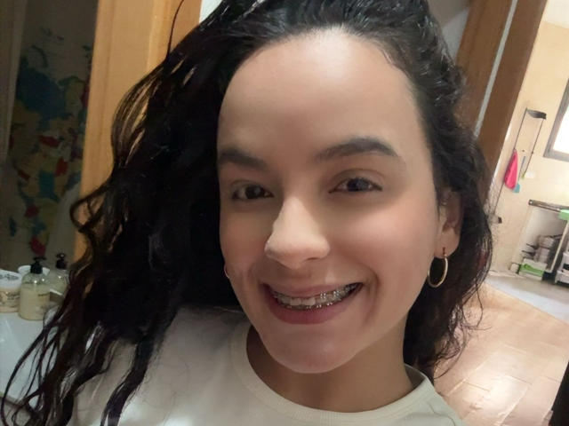MaiaPark with dark curly hair, smiling with metal braces, wearing a white shirt, and small hoop earrings, in a bathroom with wooden doors.