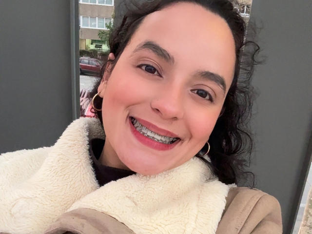 MaiaPark with curly black hair, smiling, showing metal braces, wearing a fluffy beige coat, and gold hoop earrings, looking cute and happy.