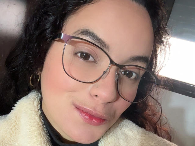 MaiaPark has curly dark hair, glasses, and pink lipstick, wearing a fluffy white jacket, with a slight smile, showing a small nose piercing on her right side.