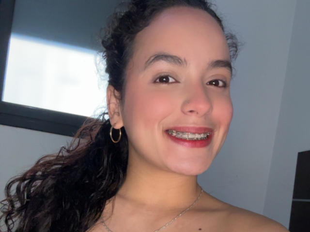 MaiaPark with curly brown hair, wearing hoop earrings and a silver necklace, smiling with metal braces, has a light skin tone and brown eyes.