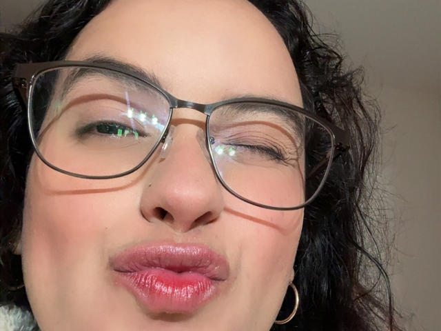 MaiaPark's face with waxy, shiny pink lips puckered in a kiss, one green eye partially closed, wearing thick black glasses, curly dark hair, and small hoop earrings.