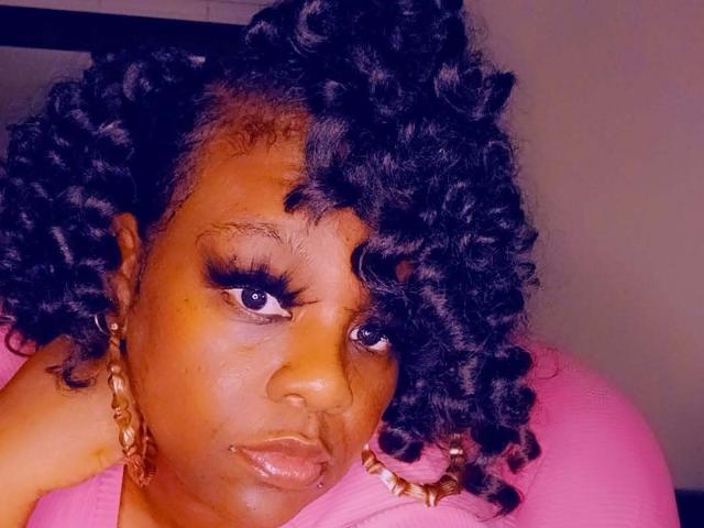 MzThundaBooty con capelli neri ricci, pelle scura, ciglia folte, orecchini a cerchio dorati e una maglietta rosa, che appare seria con un'espressione leggermente corrugata..