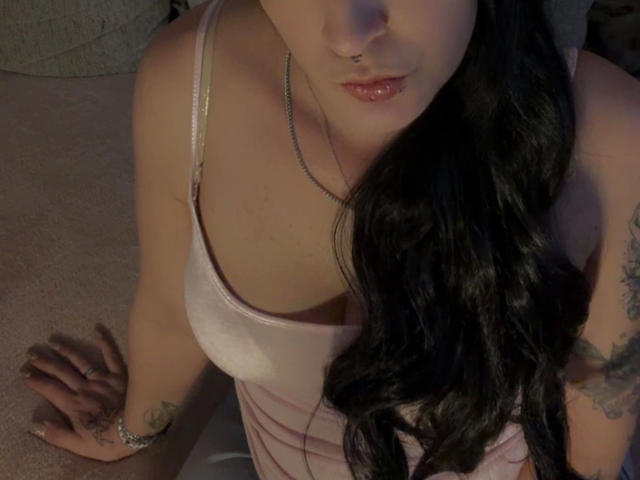 SpicyJenny - morena, com longos cabelos pretos, blusa de cetim rosa, piercing no nariz, piercing no lábio, tatuagens no braço, mão no tapete, olhando para baixo, lábios rosados levemente entreabertos, maquiagem discreta, iluminação fraca..