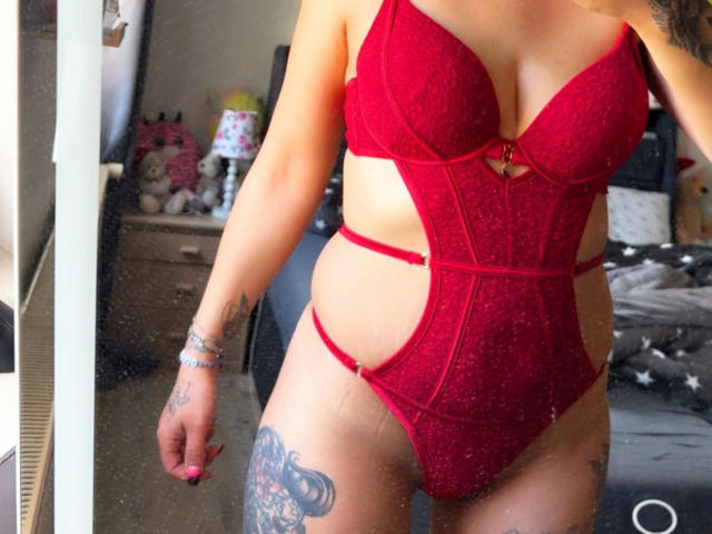 SpicyJenny, sensuelle dans une combinaison en dentelle rouge avec des découpes, dévoilant ses seins et sa cuisse tatouée, se tient devant un miroir. Elle a des ongles roses et porte un bracelet..
