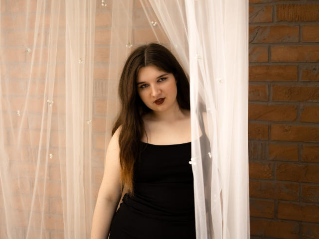 BonnieFlower with long brown hair, dark lipstick, and black dress, standing behind sheer white curtain, looking at camera, showing her bare shoulders and upper chest.