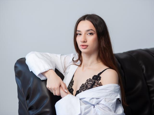 JessicaBreeze with long brown hair, wearing a white shirt off her shoulders, exposing black lace bra with her tits visible, sitting on a black leather couch.