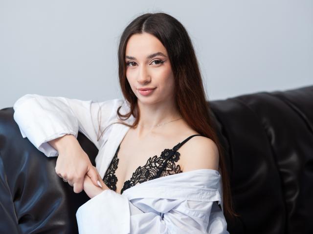 JessicaBreeze with long brown hair, wearing a white shirt off her shoulders, showing black lace bra with her tits, sitting on a black leather couch, smiling softly.