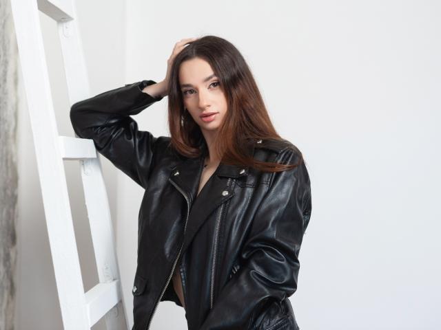JessicaBreeze with long brown hair, wearing a black leather jacket, leaning against a white ladder, hand in her hair, looking at the camera, small tits, attractive, white background.