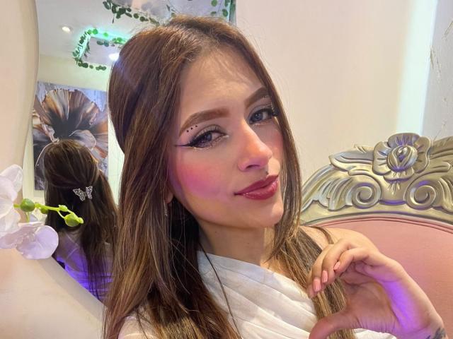 AfroditaWild with long brown hair, pink blush, and black eyeliner, smiling, wearing a white top, sitting on a pink and silver ornate chair, in a mirror reflection.
