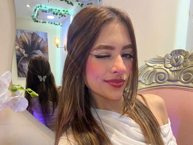 AfroditaWild with long brown hair, wearing white off-shoulder top, pink blush, black eyeliner, nose piercing, smiling, pink lipstick, butterfly hairpin, reflection in mirror, pink ornate headboard.