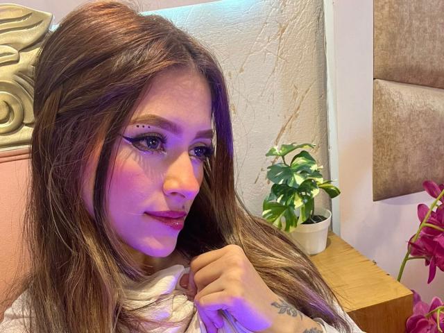AfroditaWild with long brown hair, purple eyeliner, and red lipstick, holding her hair, has a tattoo on her hand and a small plant in the background.