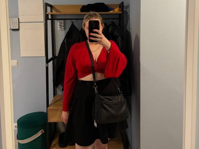 KatharinaSexy with blonde hair, red crop top, black skirt, black leather bag, holding phone, mirror selfie, showing her tits and ass, in a locker room.