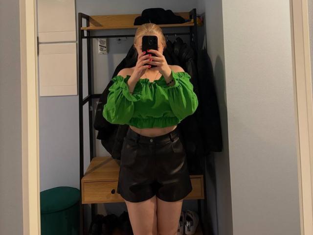 KatharinaSexy with blonde hair, green off-shoulder top, and black leather shorts, taking a mirror selfie, showing her ass and midriff, in a room with a closet.