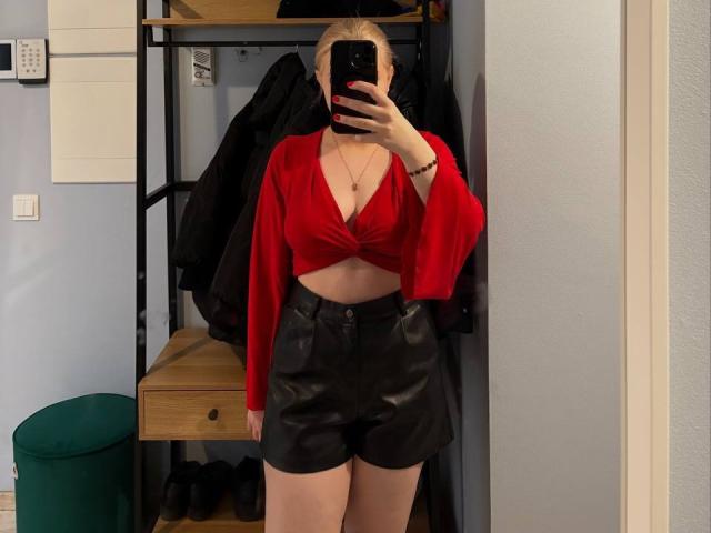 KatharinaSexy with blonde hair, red tied-up crop top showing her tits, black leather shorts, holding phone, mirror selfie, taking a picture of her reflection.
