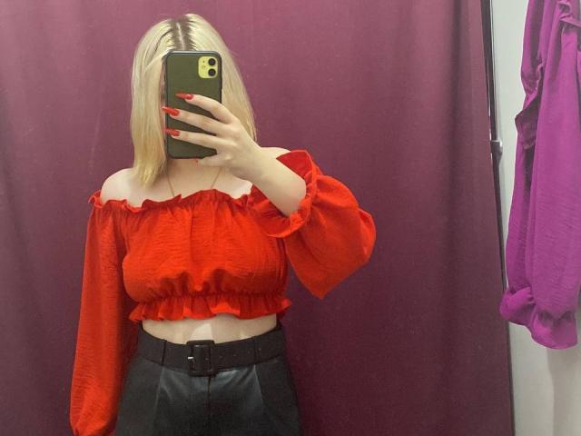 KatharinaSexy with blonde hair, red off-shoulder top, and black leather skirt, holding a phone, covering her face, red nails, visible belt, mirror selfie.