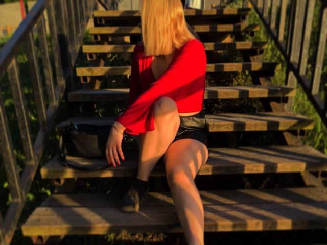 KatharinaSexy with blonde hair, red long-sleeve shirt, black leather shorts, black shoes, sitting on wooden stairs, shadow on leg, black bag on step, sunlit.