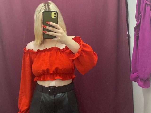 KatharinaSexy with blonde hair, red off-shoulder top, red nails, black leather skirt, holding phone in front of face, mirror selfie, visible breasts, red background.