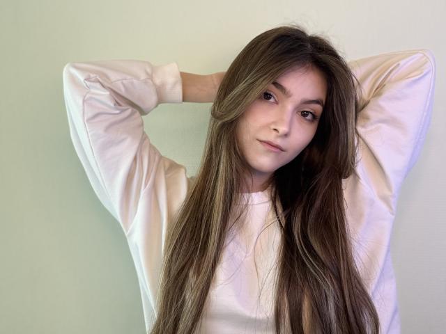 RonnaBarken with long brown hair, wearing a white shirt, arms raised, hands behind head, soft smile, light skin, subtle makeup, cute, casual, relaxed, natural light.