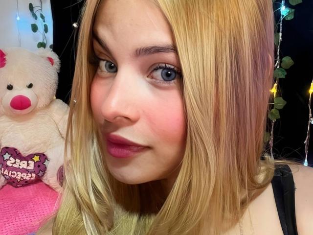 ShantalSummer has blonde hair, blue eyes, pink lipstick, and a cute face with a slight smile. Her black strap top is visible. A fluffy white teddy bear is behind her.