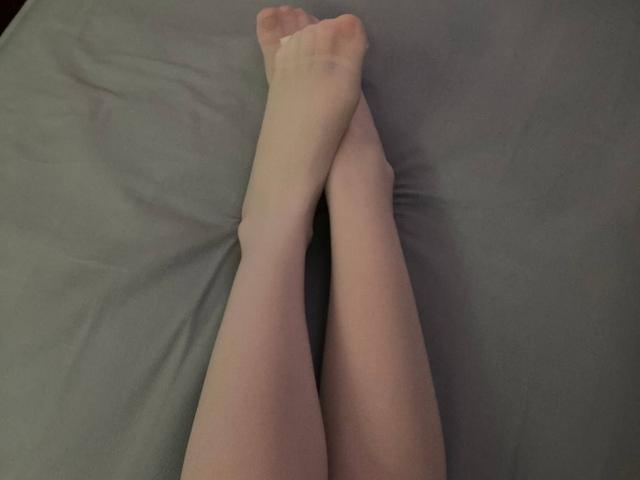 FerminaHilmer's pale legs crossed at the ankles, feet touching, lying on a gray bedsheet. Her toes are slightly pointed.