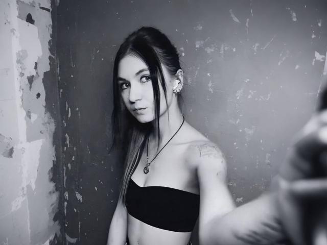 TheresaElgart with dark, straight hair, black tube top, small tattoos on shoulder, necklace, and earrings, taking a selfie, gray, slightly textured wall, serious expression, blurred arm in foreground.