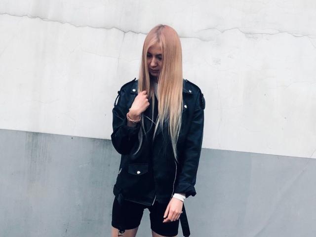 TheresaElgart has long blonde hair, wearing a black leather jacket and black shorts, with a slight smirk, standing against a white and gray wall.