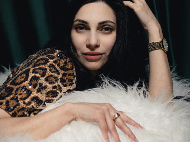 BeatriceLovely with dark hair, leopard print shirt, gold watch, and ring, laying on white fur, looking at camera with a subtle smile, hand in hair.